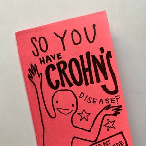 Op de afbeelding: Een roze boekje met handgeschreven tekst en illustraties in zwart. De cover luidt "SO YOU HAVE CROHN'S DISEASE?" met een tekening van een persoon en de tekst "A PATIENT WRITTEN GUIDE".