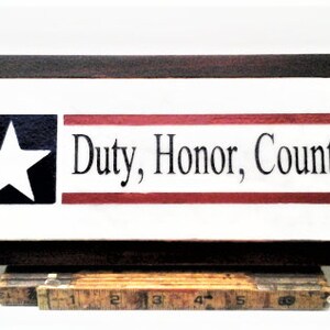 May include: A framed wooden sign with a white background and red and blue stripes. The sign features a white star on a blue background and the text "Duty, Honor, Country".