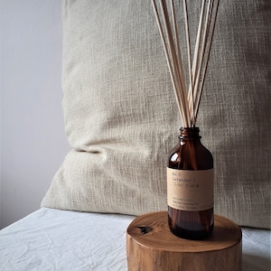 May include: A brown glass bottle with a label that reads "No. 5 Lavender + Lilac Yin & Yang" and a wooden lid. The bottle is filled with a clear liquid and has wooden sticks inserted into it. The bottle is sitting on a wooden round log.