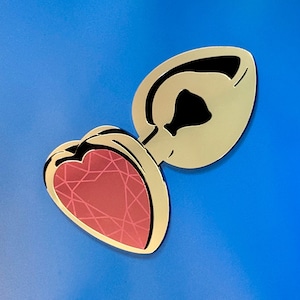 May include: A gold-colored, heart-shaped plug with a red, gem-like center and a black accent. The plug is set against a bright blue background. The design is a stylized, decorative item.