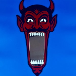 May include: A devil-shaped mirror in red and black. The devil has horns, pointed ears, and a wide open mouth with white teeth. The mirror is framed in red and set against a blue background.