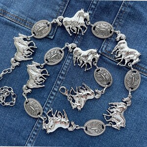 May include: Silver-toned metal belt featuring multiple horse figures and oval-shaped decorative links. The belt has a chain closure and is displayed on a denim background. The horse figures are intricately detailed.