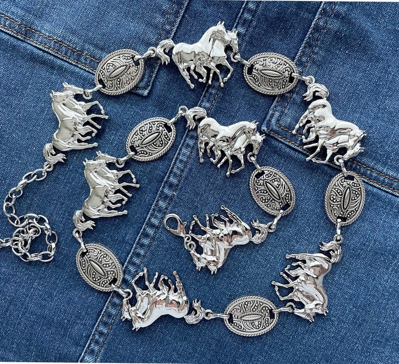 May include: Silver-toned metal belt featuring multiple horse figures and oval-shaped decorative links. The belt has a chain closure and is displayed on a denim background. The horse figures are intricately detailed.