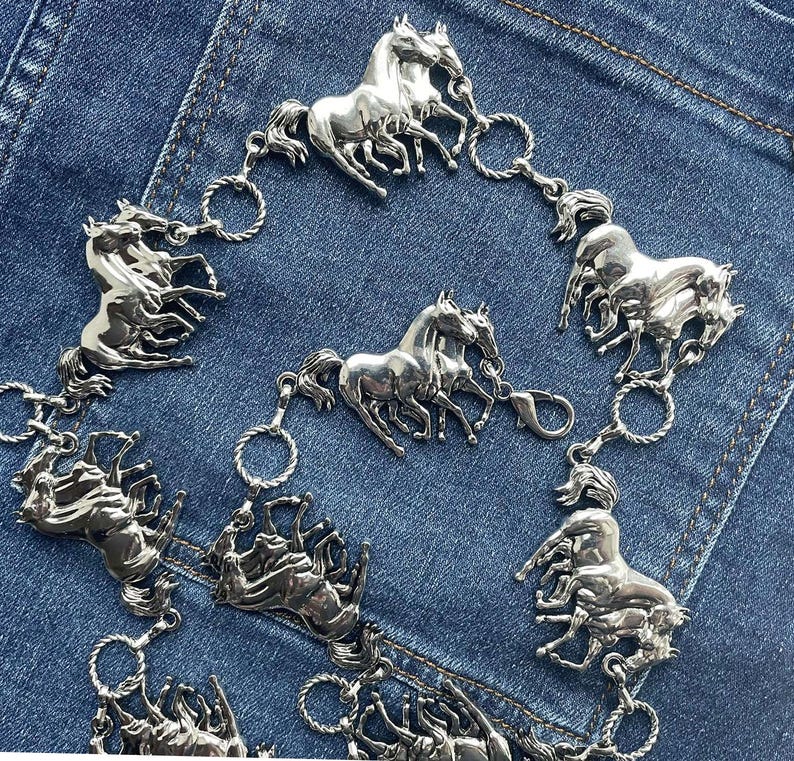 May include: Silver-toned metal belt featuring multiple horse figures. The belt is composed of several horse-shaped charms connected by small, circular links. The horses are depicted in various poses, creating a dynamic and detailed design. The belt is set against a denim background.