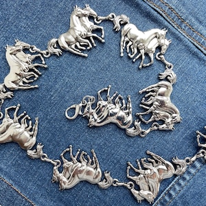 May include: Silver-toned horse-themed belt. The belt features multiple detailed horse figures linked together, with a clasp closure. The horses are arranged in a circular pattern. The belt is displayed on a blue denim background.