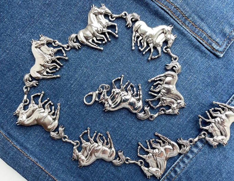 May include: Silver-toned horse-themed belt. The belt features multiple detailed horse figures linked together, with a clasp closure. The horses are arranged in a circular pattern. The belt is displayed on a blue denim background.