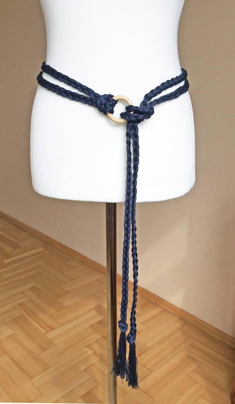 Peut inclure: Une ceinture en corde tress&eacute;e bleu marine avec un anneau en bois et des glands. La ceinture est nou&eacute;e autour d'un mannequin blanc.