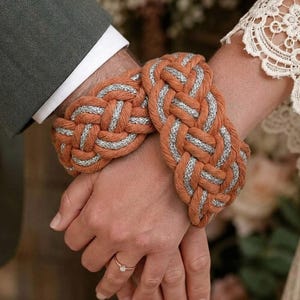 May include: Two braided wristbands featuring a thick, interwoven pattern of burnt orange cord and shimmering silver metallic thread. The bracelets are worn on two wrists held together, with one wrist showing a thin gold ring with a small clear stone.