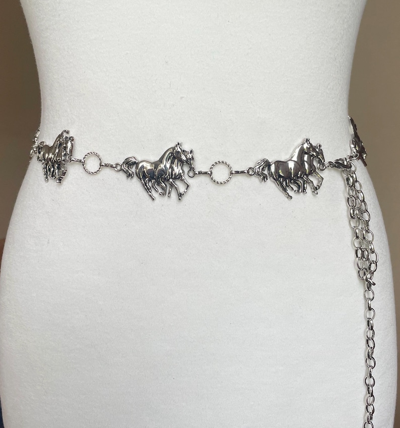 May include: Silver-toned metal belt featuring horse figures. The belt has a chain with horse-shaped charms and a dangling chain. The horse charms are evenly spaced along the belt, creating a Western-inspired accessory.
