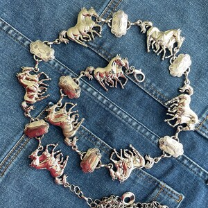 May include: Silver-toned belt featuring multiple horse figures and decorative floral-shaped links. The belt is composed of several horse-shaped charms connected by small links and floral-shaped accents. The belt is displayed on a blue denim background.