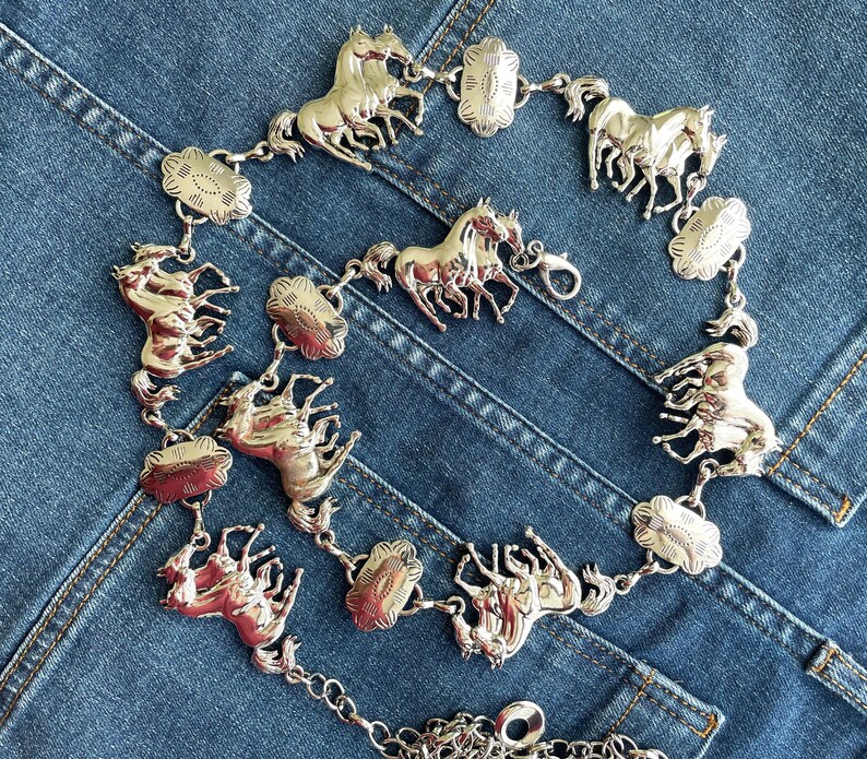 May include: Silver-toned belt featuring multiple horse figures and decorative floral-shaped links. The belt is composed of several horse-shaped charms connected by small links and floral-shaped accents. The belt is displayed on a blue denim background.