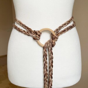 Peut inclure: Une ceinture en corde tress&eacute;e marron et blanche avec un anneau en bois. La ceinture est nou&eacute;e &agrave; l'avant et les extr&eacute;mit&eacute;s de la corde pendent.