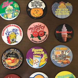 May include: Twelve round, colourful, vintage McDonald's buttons with various themes, including Happy Meal, McRib, Banana Shakes, and Pineapple Pies. The buttons feature cartoon characters, food items, and slogans.