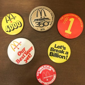 Peut inclure: Ensemble de badges et d'une pièce commémorative McDonald's vintage. Les badges sont jaunes, rouges et blancs, avec des textes tels que "4000", "Our Best Year yet!" et "Let's Break a Billion". La pièce commémore le 350e magasin.