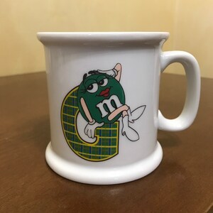 May include: White ceramic mug featuring a green M&M's character with a red lipstick smile, reclining on a yellow and blue plaid letter "G". The mug has a wide base and a curved handle.
