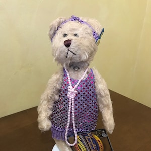 Peut inclure: Un ours en peluche brun clair portant une robe pailletée violette avec des franges roses et un collier de perles roses. L'ours porte un bandeau violet avec un bouton doré. L'ours est debout sur un piédestal blanc et porte une étiquette qui indique "The Charles Taylor Collection - Precious Ballerina Bears".