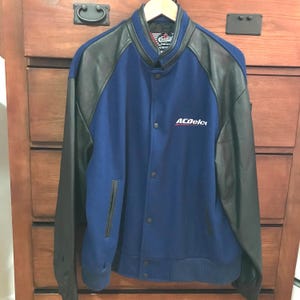 Peut inclure: Une veste de baseball bleu et noire avec des manches en cuir. La veste a une fermeture à pression et un logo brodé blanc qui dit "ACDelco".