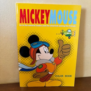 Peut inclure: Un livre de coloriage jaune avec Mickey Mouse portant une tenue de pilote et faisant un pouce en l'air. Le livre contient le texte "MICKEY MOUSE" et "he is the natural leader and the smartest among his friends." Le livre contient également le texte "COLOR BOOK".
