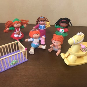 Peut inclure: Un ensemble de sept poupées Cabbage Patch Kids et d'accessoires, comprenant un berceau, un cheval à bascule et une poupée tenant un ours en peluche. Les poupées portent toutes des tenues différentes et ont des couleurs de cheveux différentes.