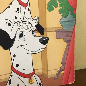 以下が含まれることがあります： 2匹のダルメシアンの犬の漫画イラスト。大きな犬は頭を横に傾けて立っており、小さな犬がその頭の上に座っています。両方の犬は赤い首輪に金のタグが付いています。