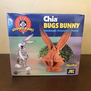 May include: A box for a Chia Pet featuring Bugs Bunny from Looney Tunes. The box shows a brown ceramic planter shaped like Bugs Bunny's head with green chia growing out of it. The box also shows a cartoon image of Bugs Bunny holding a carrot.