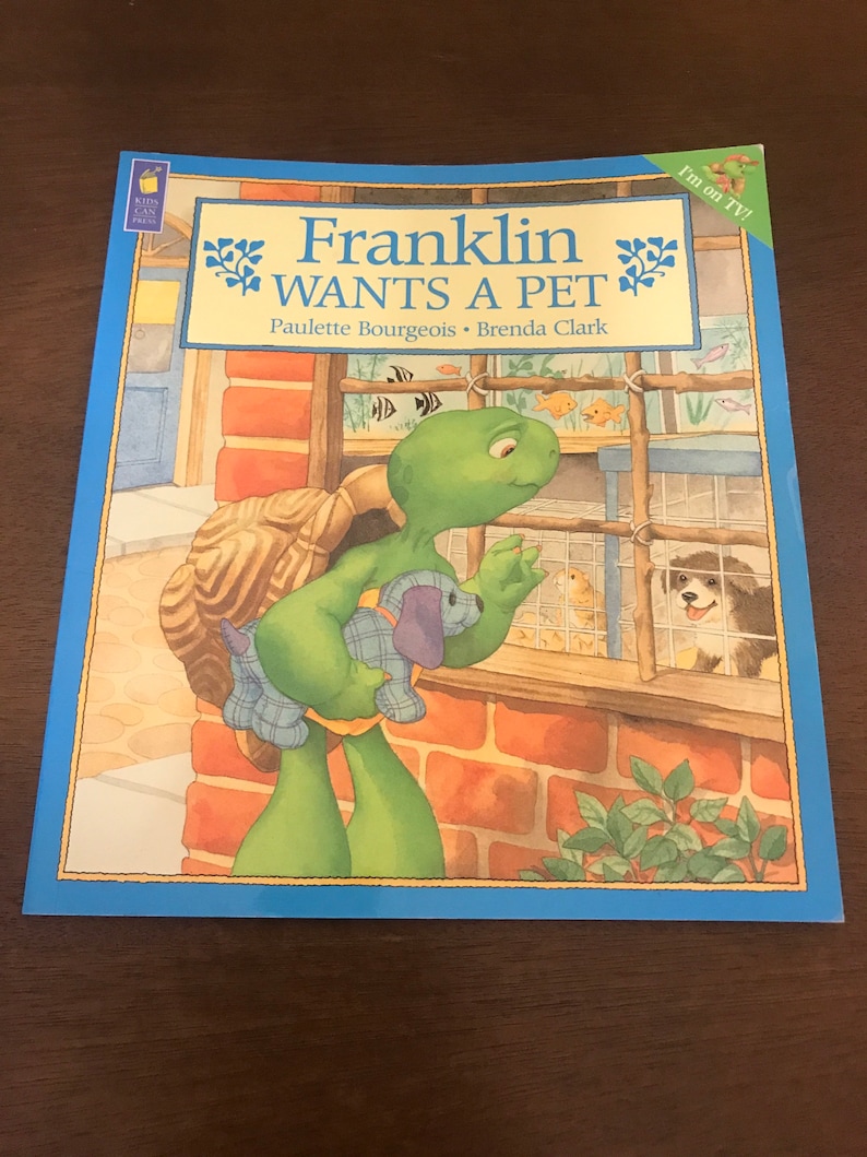 Peut inclure: Une tortue verte nomm&eacute;e Franklin tenant un chiot en peluche, regardant un chiot dans une cage. La couverture du livre indique "Franklin veut un animal de compagnie" et comporte le texte "Je suis &agrave; la t&eacute;l&eacute; !" dans un encadr&eacute; jaune.