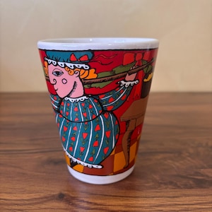May include: A ceramic shot glass with a colorful illustration of a person in a teal dress with red heart accents. The background is red with brown furniture. The rim of the glass is white.