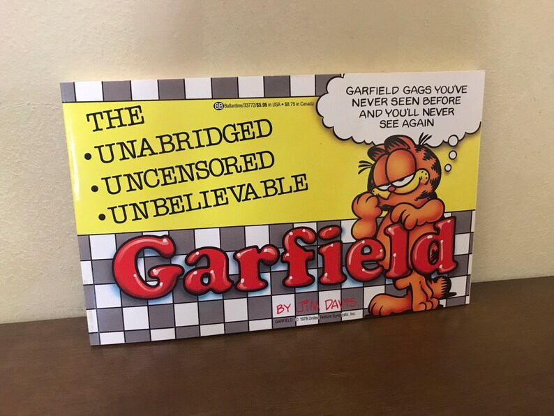May include: A paperback book titled The Unabridged Uncensored Unbelievable Garfield by Jim Davis. The cover features a yellow background with a black and white checkered pattern at the bottom, an illustration of Garfield, and a speech bubble with text.