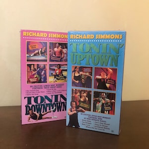 May include: Two VHS tapes featuring Richard Simmons's exercise videos. The tapes are titled "Tonin' Uptown" and "Tonin' Downtown". The tapes feature images of people exercising and the names of the exercises they are doing. The tapes are pink and blue.