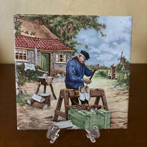 Peut inclure: Un carreau vintage représentant un homme en chemise bleue et casquette noire sculptant des sabots en bois dans un cadre rural. La scène comprend une petite maison avec un toit de chaume, un moulin à vent et une clôture en bois.