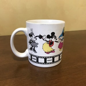 May include: White ceramic mug with a black and white image of Mickey Mouse and Minnie Mouse from different eras. The mug has a black film strip design with the years 1928 and 1937 printed on it.