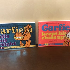 May include: Two Garfield comic books by Jim Davis. The left book is titled Garfield Pulls His Weight, His 26th Book, with a dark blue cover. The right book is titled Garfield Keeps His Chins Up, His 23rd Book, with a bright orange cover and Garfield illustration.