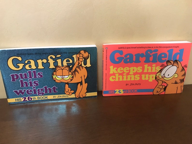 May include: Two Garfield comic books by Jim Davis. The left book is titled Garfield Pulls His Weight, His 26th Book, with a dark blue cover. The right book is titled Garfield Keeps His Chins Up, His 23rd Book, with a bright orange cover and Garfield illustration.