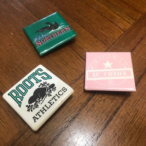 Peut inclure: Trois épingles carrées avec des motifs différents. La première épingle est verte avec un huard noir et le texte "NORTHERN" en rose. La deuxième épingle est blanche avec un castor noir et le texte "ROOTS ATHLETICS" en bleu. La troisième épingle est rose avec une étoile blanche et le texte "ORIGINAL ALL COTTON (R)" en noir.