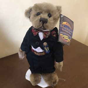 May include: A brown teddy bear wearing a black tuxedo with a red bow tie and a gold pin that says "2000 Century". The bear is standing on a white pedestal and has a tag that says "Les Cousins Park Bears -  Danny Button Bear -  Edition du Millenaire 2000".