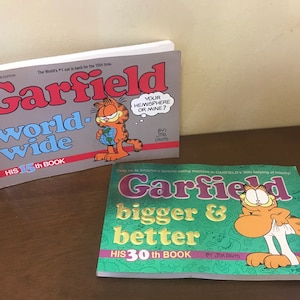 May include: Two Garfield comic books. The first book is titled Garfield worldwide, his 15th book, featuring an orange cat holding a globe. The second book is titled Garfield bigger & better, his 30th book, featuring an orange cat pulling up white underwear.