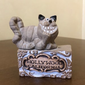 May include: A ceramic figurine of a grinning cat with black ears and eyes, sitting on a ceramic block with the words "HOLLYWOOD CALIFORNIA" inscribed on it.
