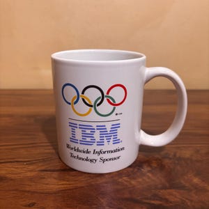 May include: White ceramic mug with the Olympic rings in blue, black, red, yellow, and green. The IBM logo is below the rings, with the text "Worldwide Information Technology Sponsor". The mug has a curved handle.