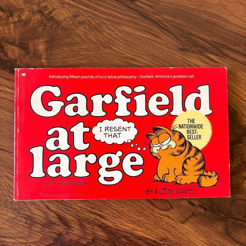 May include: A red paperback book titled Garfield at Large by Jim Davis. The cover features a cartoon orange tabby cat, a speech bubble with the text I RESENT THAT, and a yellow starburst graphic stating THE NATIONWIDE BEST-SELLER.