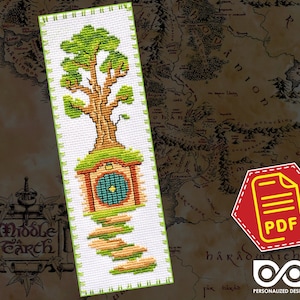 May include: Cross stitch pattern for a bookmark featuring a green tree with a brown trunk and a small house with a blue door. The house is surrounded by a green path. The pattern is on white fabric.