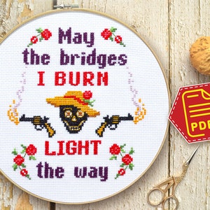 May include: A cross-stitch embroidery hoop with the text "May the bridges I BURN LIGHT the way." The design features a skull wearing a hat, two guns, and floral accents. A PDF icon and gold scissors are also visible.