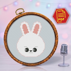 May include: A cross-stitch embroidery hoop featuring a white bunny design with pink inner ears and black eyes and mouth. The embroidery is set in a wooden hoop with a brass clasp. A red PDF icon is in the upper right corner.