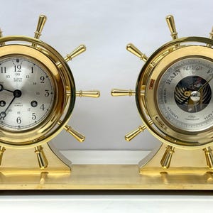 May include: A brass ship's wheel clock and barometer set. The clock face has black Roman numeral hour markers and black hands. The barometer face has a white background with black markings and a silver needle.