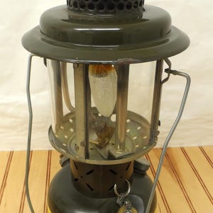 May include: A green metal camping lantern with a glass globe and a yellow warning label. The lantern has a metal handle and a metal fuel tank.