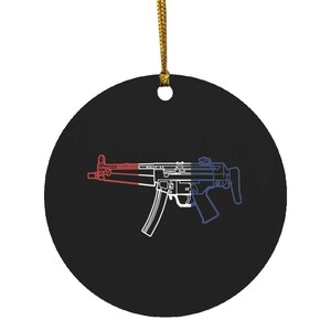 May include: A round black ornament featuring a white line art illustration of a submachine gun. The firearm design is divided into three vertical color sections of red, white, and blue, and the ornament hangs from a gold-colored string.