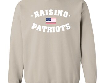 May include: A sand-colored crewneck sweatshirt featuring the white text RAISING PATRIOTS in an arched layout. Between the two words is a small, rectangular graphic of the United States flag. There is a small white star on each side of the text.