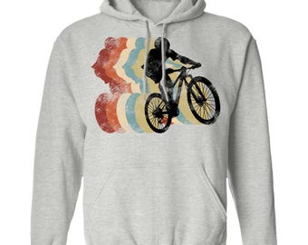 以下が含まれることがあります： マウンテンバイクに乗るサイクリストのグラフィックが描かれたライトグレーのフード付きスウェットシャツ。赤、オレンジ、黄色、青の色合いの、かすれた多色の円形パターンが、黒いサイクリストのシルエットの背後にデザインされています。
