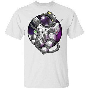 May include: White t-shirt with a graphic of an astronaut floating in space. The astronaut is wearing a white spacesuit with a purple and black galaxy background.