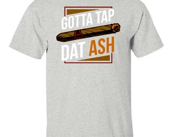 May include: A light gray short-sleeve t-shirt featuring a graphic of a brown cigar centered between the white text "GOTTA TAP" and "DAT ASH" in orange, all enclosed within a thin dark red rectangular border on the front of the garment.