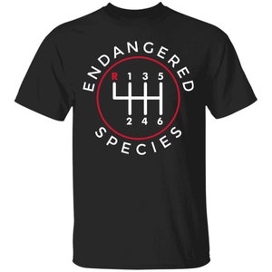 May include: Black t-shirt with a white and red graphic. The graphic features a gear shift pattern inside a red circle, with the words "ENDANGERED SPECIES" around the circle. The gear shift shows R, 1, 3, 5, 2, 4, and 6.